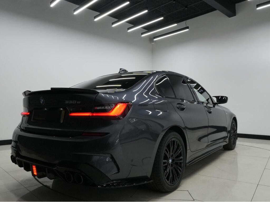 A sleek, dark-colored BMW 330e parked indoors with bright, linear ceiling lights reflecting on its polished surface. The rear of the car features illuminated taillights and visible exhaust pipes.