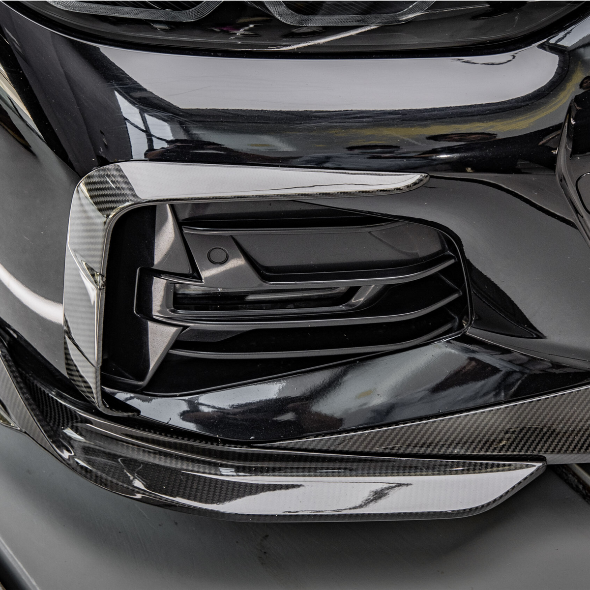 A black car with a carbon fiber front bumper detail is parked in a garage, reflecting nearby objects and lights on its glossy surface.