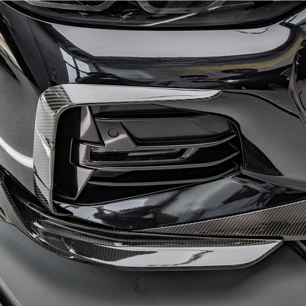 A black car with a carbon fiber front bumper detail is parked in a garage, reflecting nearby objects and lights on its glossy surface.