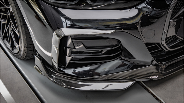 A black car with a carbon fiber front bumper detail is parked in a garage, reflecting nearby objects and lights on its glossy surface.