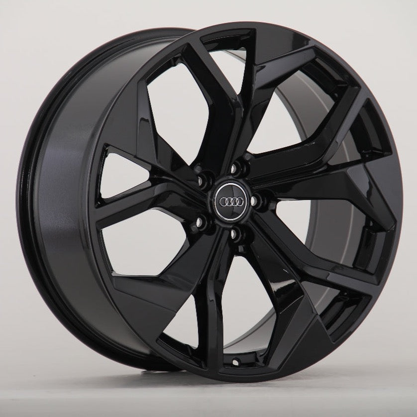 A glossy black alloy wheel with a seven-spoke design prominently displays an Audi logo at its center, positioned against a plain, neutral background.
