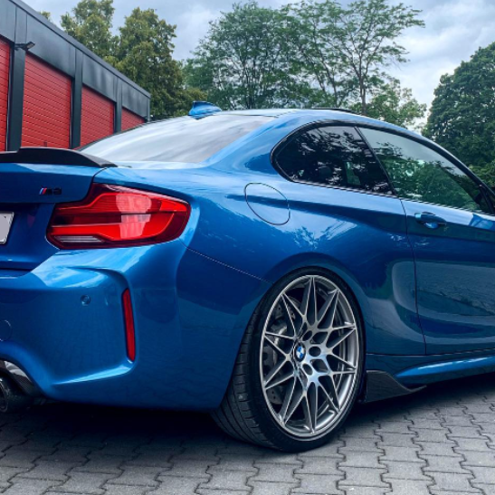 A blue sports car, parked on a paved area, features sleek alloy wheels and a spoiler. It's positioned in front of a building with red panels, surrounded by trees.