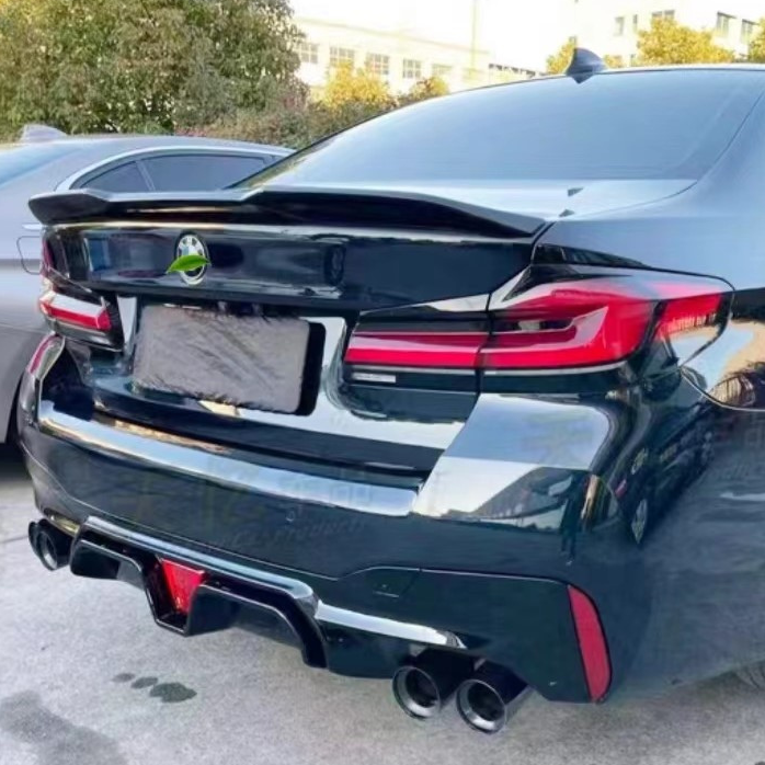 A black car parked in a lot has a prominent rear spoiler, dual exhaust pipes, and tinted windows. Next to it is a gray car. Trees and a building are in the background.
