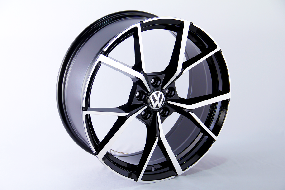 A black and silver alloy wheel stands upright, showcasing a geometric design with a central Volkswagen logo, set against a plain white background.