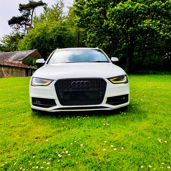 A white Audi car is parked on a grassy field, surrounded by lush green trees and a rustic building in the background.