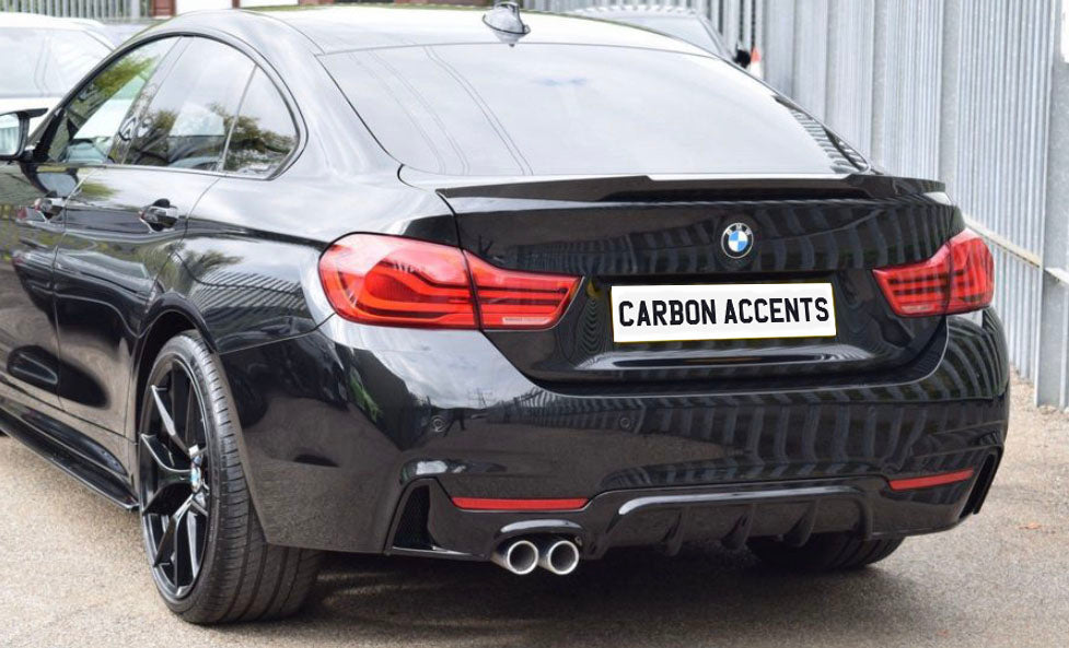 A black BMW car is parked on a street beside a metal fence. The license plate reads "CARBON ACCENTS," and it features dual exhaust pipes and red tail lights.