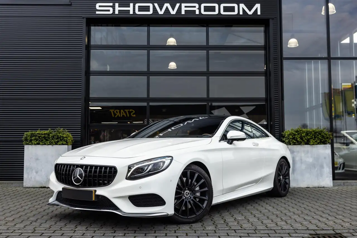 A white Mercedes-Benz car is parked outside a building with "SHOWROOM" written above a large glass door. Two potted plants flank the car on a paved surface.