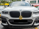 A white BMW SUV is parked, displaying a front grille with a badge. It's flanked by other cars on a paved area, with a building and trees in the background.