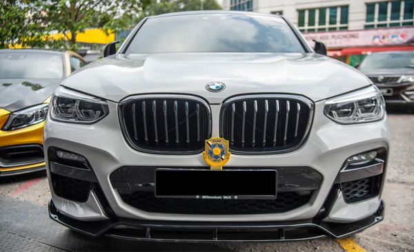 A white BMW SUV is parked, displaying a front grille with a badge. It's flanked by other cars on a paved area, with a building and trees in the background.