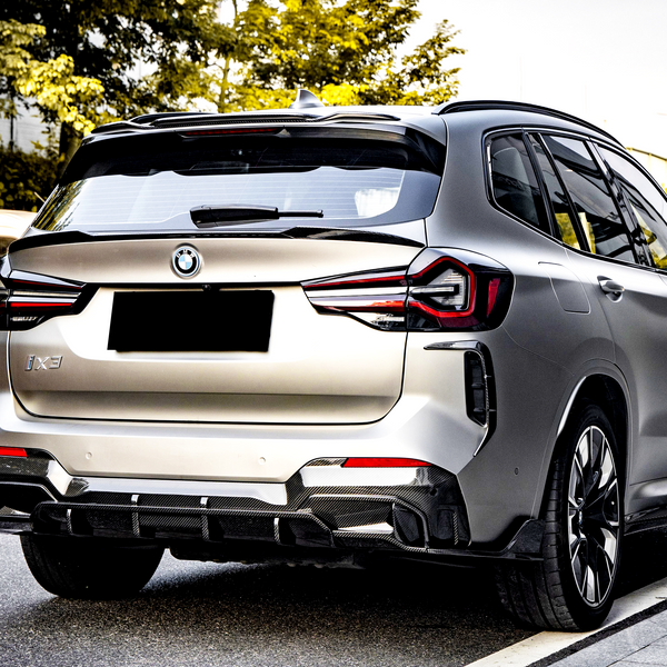 The object is a silver BMW iX3 SUV parked on a city street. It features distinctive taillights and a black license plate. Trees and other vehicles are nearby.