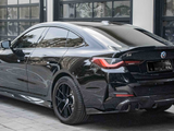 A black BMW i4 parked on a cobblestone driveway outside a modern building with large windows. The car's design features sleek lines and tinted windows, with badges displaying 