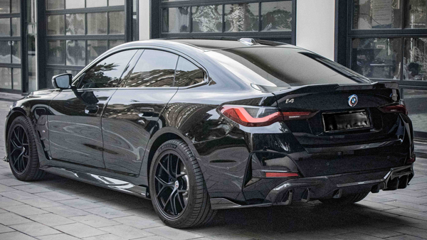 A black BMW i4 parked on a cobblestone driveway outside a modern building with large windows. The car's design features sleek lines and tinted windows, with badges displaying "BMW," "i4," and "M50".