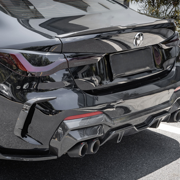 A black sports car is parked on a street, featuring a sleek rear design, quad exhaust pipes, and a BMW logo. Surrounding foliage lines a nearby concrete boundary.