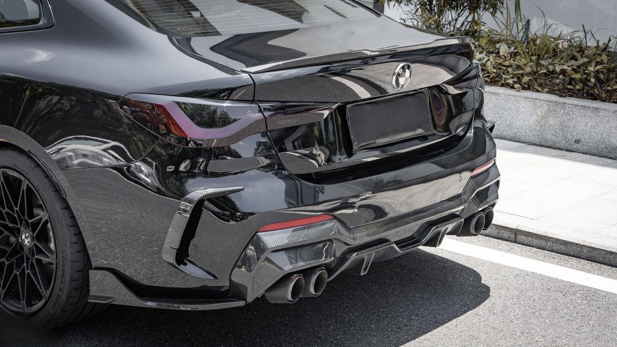 A black sports car is parked on a street, featuring a sleek rear design, quad exhaust pipes, and a BMW logo. Surrounding foliage lines a nearby concrete boundary.