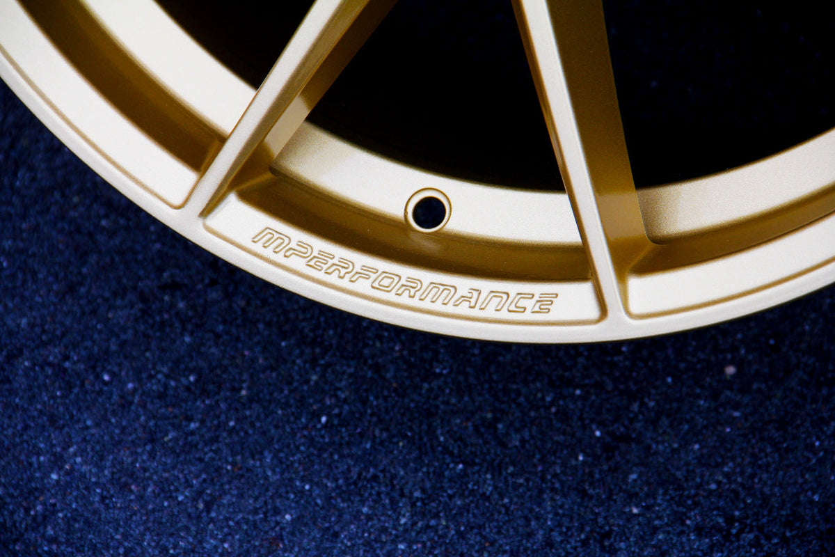 A close-up of a gold alloy wheel rim featuring embossed text "MPERFORMANCE," set against a textured, dark blue surface.