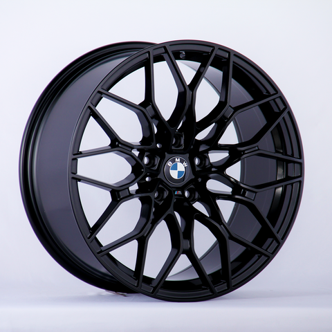 A black, intricately designed BMW alloy wheel is propped against a plain white background, featuring the BMW logo prominently at the center.