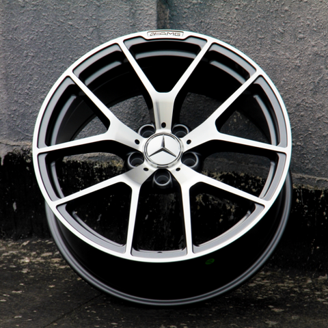 A metallic alloy wheel with a star emblem and "AMG" logo is standing upright against a textured gray wall, positioned on a dark concrete surface.