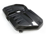 A carbon fiber engine cover with a sleek, angular design lying on a plain white surface. The cover has a central circular opening and raised geometric patterns for added texture.