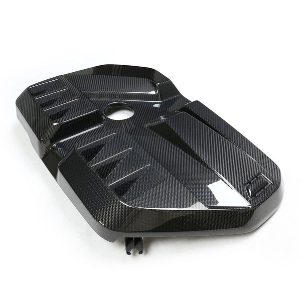 A carbon fiber engine cover with a sleek, angular design lying on a plain white surface. The cover has a central circular opening and raised geometric patterns for added texture.