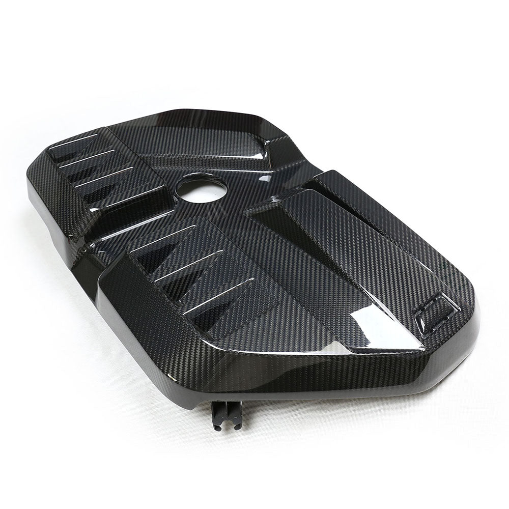 A carbon fiber engine cover with a sleek, angular design lying on a plain white surface. The cover has a central circular opening and raised geometric patterns for added texture.