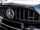 A black car grille features a prominent Mercedes-Benz logo and AMG badge. The grille has vertical slats, reflecting sunlight. Parked vehicles and a building are faintly visible in the background.