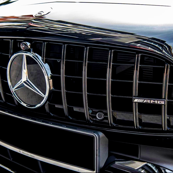A black car grille features a prominent Mercedes-Benz logo and AMG badge. The grille has vertical slats, reflecting sunlight. Parked vehicles and a building are faintly visible in the background.