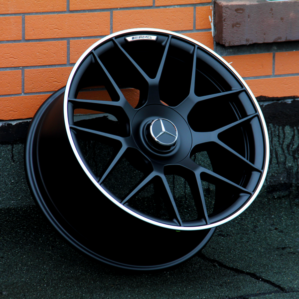 A black Mercedes-Benz AMG wheel rim leans against an orange brick wall on a dark concrete surface. The central emblem features the Mercedes logo.