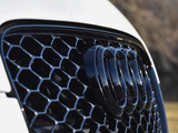 The car's front grille features a glossy black mesh pattern with four intersecting rings emblem; blurred trees and grass are in the background.