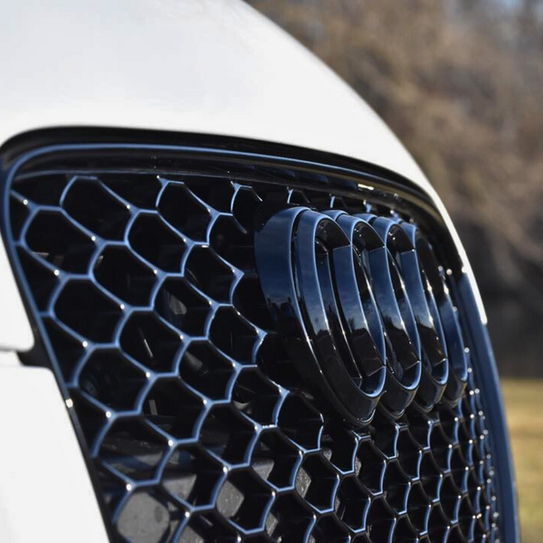 The car's front grille features a glossy black mesh pattern with four intersecting rings emblem; blurred trees and grass are in the background.