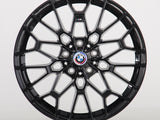The image shows a black alloy wheel with a complex geometric design, featuring a blue, white, and red BMW logo at the center. The wheel is set against a plain white background.