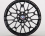 The image shows a black alloy wheel with a complex geometric design, featuring a blue, white, and red BMW logo at the center. The wheel is set against a plain white background.