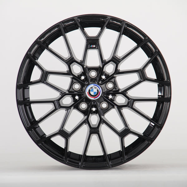 The image shows a black alloy wheel with a complex geometric design, featuring a blue, white, and red BMW logo at the center. The wheel is set against a plain white background.