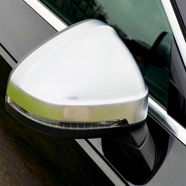 A silver car side mirror is affixed to a black vehicle, reflecting surrounding greenery. The mirror features an integrated turn signal beneath its polished surface.