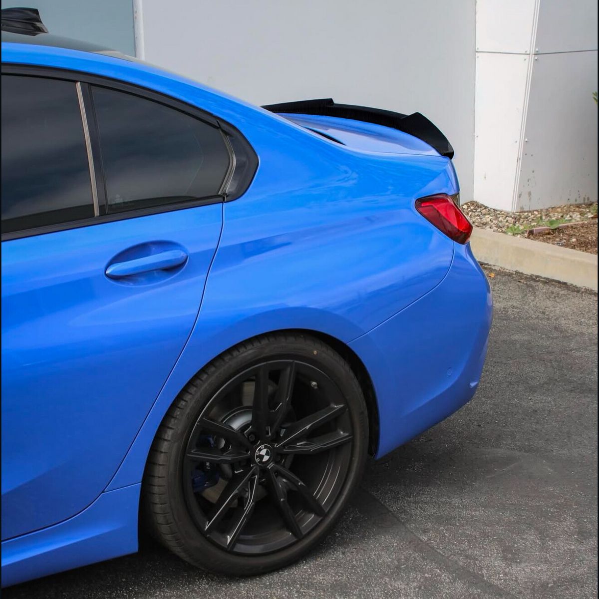A blue car is parked on an asphalt surface, featuring black alloy wheels and a rear spoiler. The vehicle is positioned near a white building wall with some landscaping nearby.