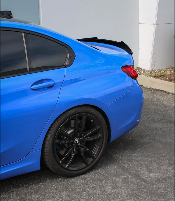 A blue car is parked on an asphalt surface, featuring black alloy wheels and a rear spoiler. The vehicle is positioned near a white building wall with some landscaping nearby.