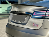 A car's rear with a smooth, metallic finish displays a Tesla logo and distinct linear taillights. The environment is an outdoor pavement area, with another vehicle partially visible.