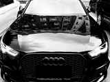 A black Audi car is parked in a lot, surrounded by other vehicles. Its hood reflects the windows of a tall building, and 