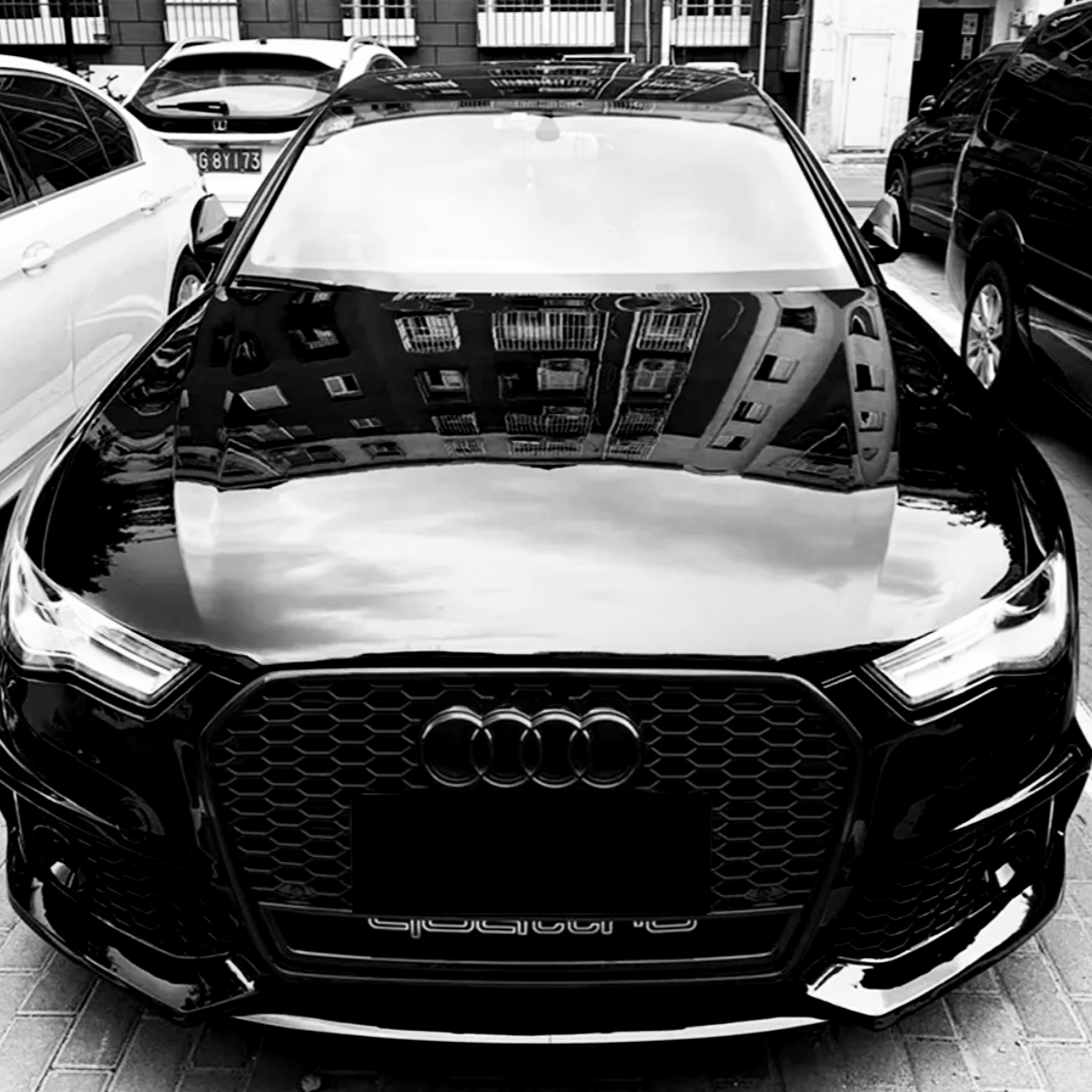 A black Audi car is parked in a lot, surrounded by other vehicles. Its hood reflects the windows of a tall building, and "quattro" is written below the grille.
