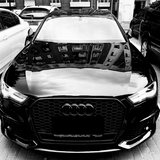 A black Audi car is parked in a lot, surrounded by other vehicles. Its hood reflects the windows of a tall building, and "quattro" is written below the grille.