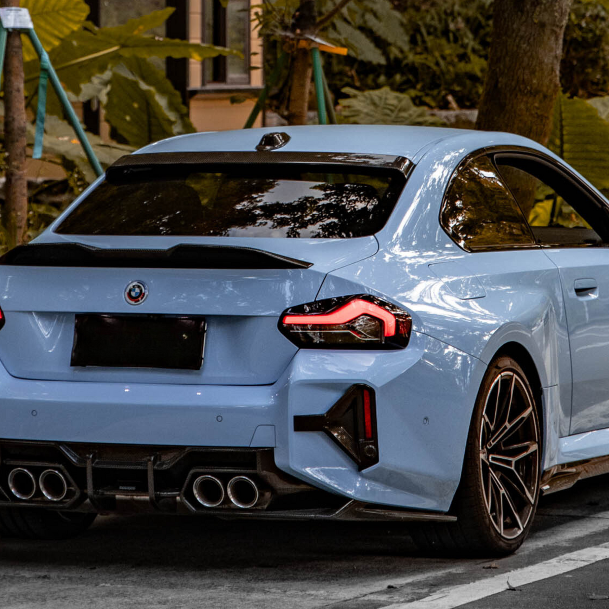 A blue sports car with distinctive tail lights and dual exhausts is parked on a road, surrounded by lush green vegetation and a staircase in the background.