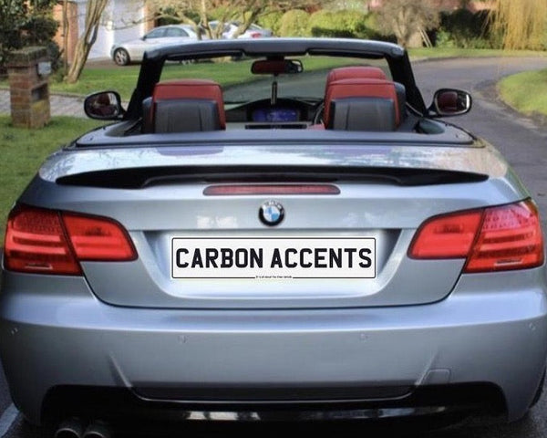 Convertible silver car with red interior parked on a residential street. License plate reads "CARBON ACCENTS." Brick mailbox and house in the background surrounded by trees and grass.