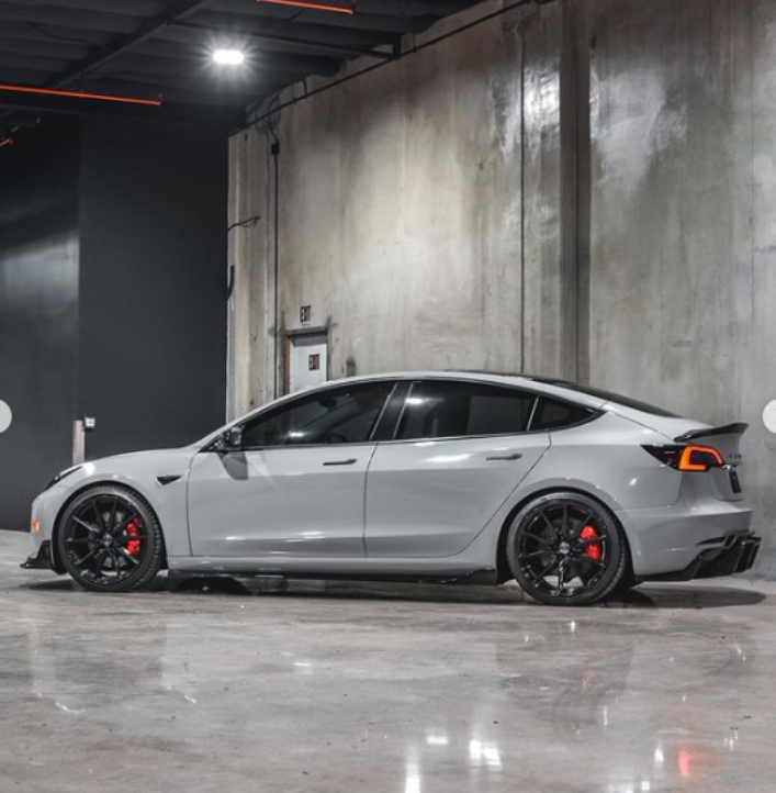 A gray sports car is parked in a sleek, industrial garage with concrete walls and polished floors. It has black wheels and red brake calipers.