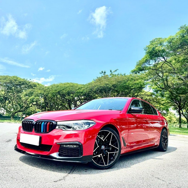 A red car is parked on a paved surface under a clear sky, surrounded by lush green trees in the background.