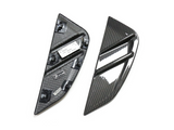 Two carbon fiber car trim pieces lie on a plain surface; one shows a glossy, ribbed exterior, while the other reveals a textured interior with mounting clips and labels.