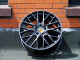 A black alloy wheel with a yellow Porsche emblem, leaning against an orange brick wall, casting a shadow on a textured ground surface.