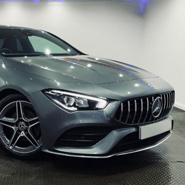 A sleek, silver luxury car is parked on a shiny floor in a brightly lit showroom, featuring a prominent front grille and stylish alloy wheels.