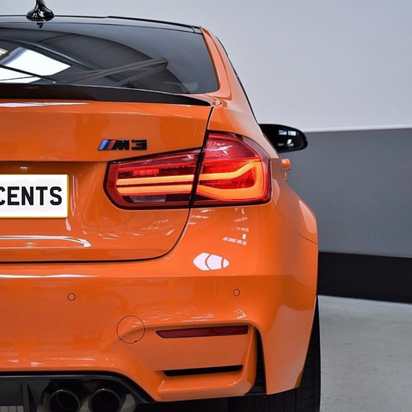 Orange BMW car with illuminated taillights is parked in a garage. The license plate reads "CARBON ACCENTS," and the car displays an "M3" badge.