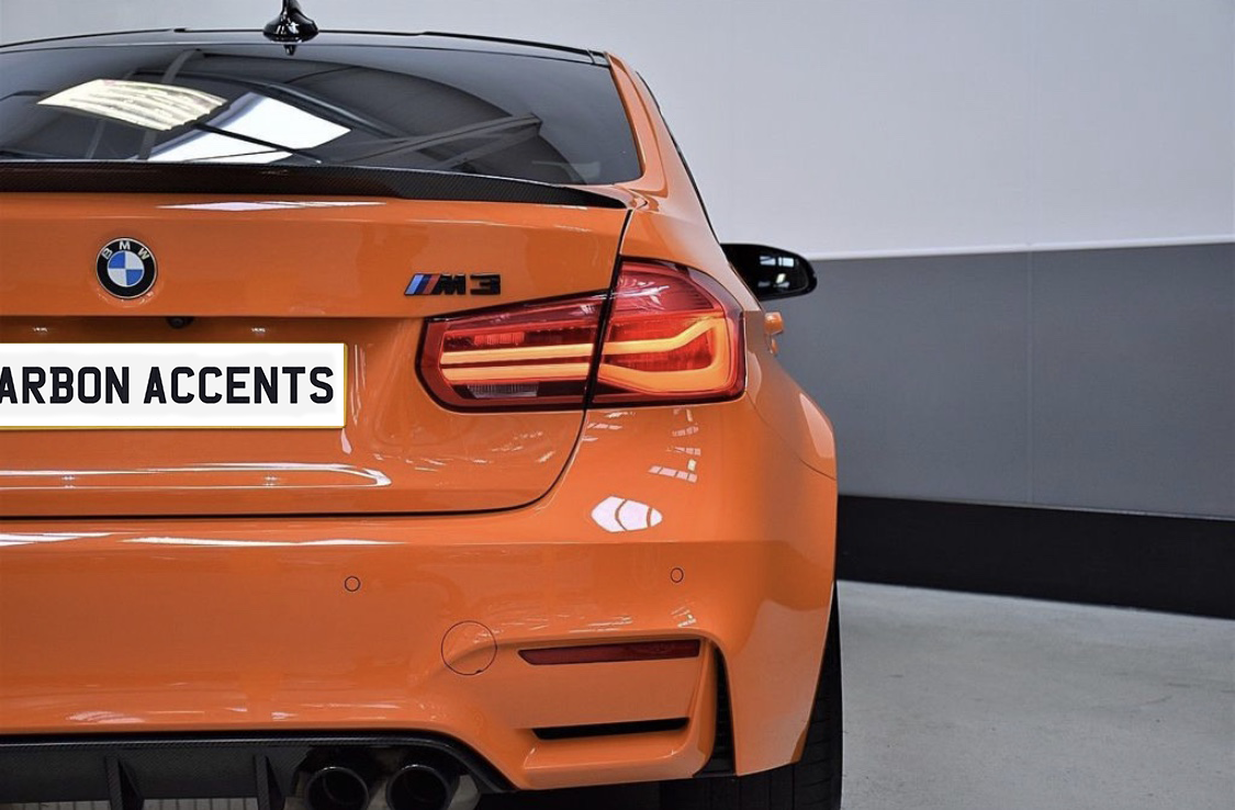 Orange BMW car with illuminated taillights is parked in a garage. The license plate reads "CARBON ACCENTS," and the car displays an "M3" badge.