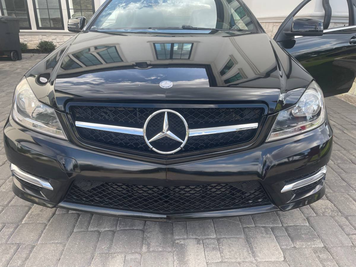 A black Mercedes-Benz car is parked on a stone driveway outside a building. The car's front grill and emblem are prominently visible, with the driver's door slightly open.