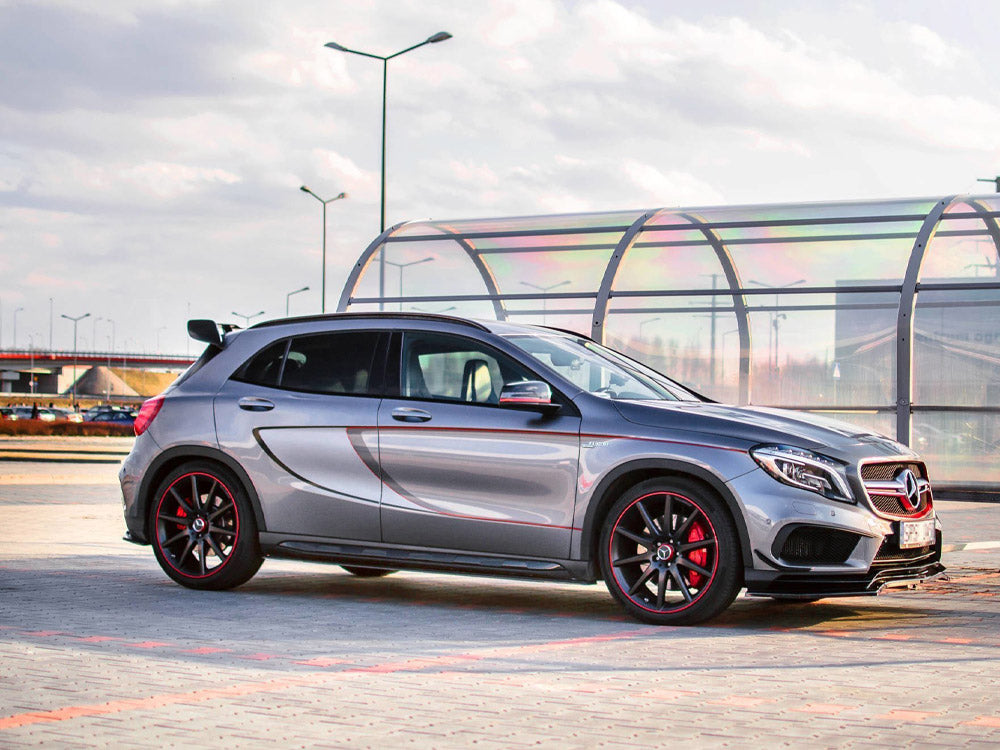 A silver hatchback car with red-accented black wheels is parked on a paved lot. The vehicle features a sleek design. The background has a glass-covered walkway and lamp posts.
